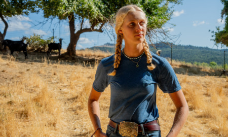 Goats - A person with two blonde braids wearing a blue t-shirt, jeans and a large belt buckle