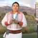 Native indigenous language: Man with black hair pulled back wearing native clothing with re detailing on white shirt stands in desert landscape i front of two adults holding signs