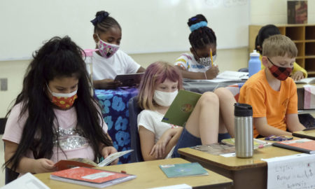 School funding addresses summer program disparities: Six masked elementary-age children sit at two rows of desks reading and writing