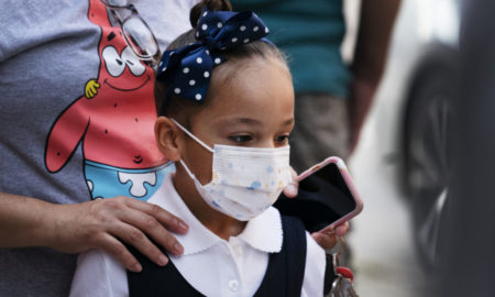 delta variant afterschool: young Elementary-age Black girl with black hair in top bun with large navy bow wearing dark navy and white top and face mask