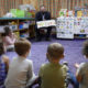 Build Back Better Child Tax Credit: Young children sit in circle on purple carpet listening to adult man in suit showing them posters