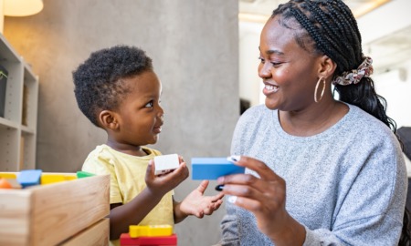 Black women are still holding up the child care industry: Black woman with long hair plays blocks with young black boy in child care setting