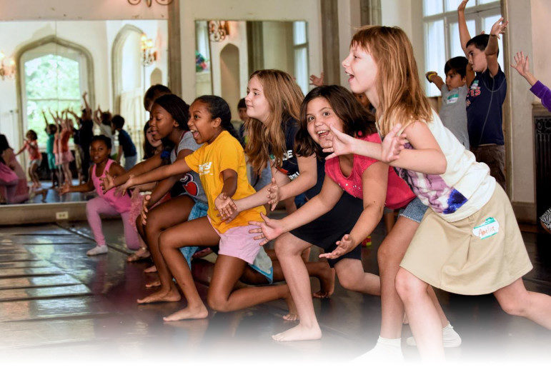 Health in OST: A dozen plus young children laughing while doing dance exercises in a dance studio reflected in full wall mirror.