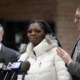 Lawsuit; Illinois agency wrongfully imprisoned children: young black woman in white coat talking into microphones with men in suits at her side