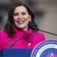 Michigan Gov. Whitmer calls for prek for all 4 year-olds: brunette woman in pink jacket smiling at podium with microphones