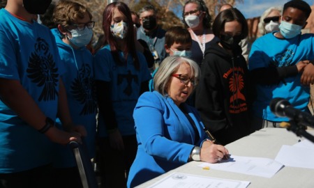 New Mexico offers equal pay to Native American teachers: older woman surrounded by people in matching blue Native logo shirts signing a document