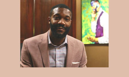 Birmingham: Smiling man with short dark hair, beard, mustache in beige jacket, checked shirt in office with painting, bookcase behind him.