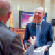 CYFD: Smiling man with red beard talks to other men in hallway, all wearing business suits.