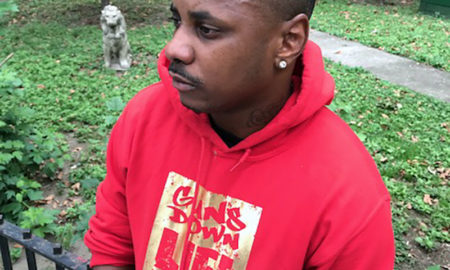 Guns Down Life Up: Man in red hoodie with words guns down life up on it looks to left, standing outside in front of apartment buildings