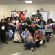 girls: Big group of smiling young women pose with cards on a string; smiling woman in gray sweater, jeans, sneakers poses in front of them with jazz hands