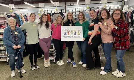 Foster care foundation: Nine women stand posing in a line smiling at the camera. Behind them are racks of hanging clothing. The two women in the middle are holding a sign featuring a graphic of children that reads "Foster Care Support www.fostercares.org"