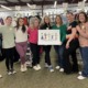 Foster care foundation: Nine women stand posing in a line smiling at the camera. Behind them are racks of hanging clothing. The two women in the middle are holding a sign featuring a graphic of children that reads "Foster Care Support www.fostercares.org"