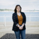 Youth homelessness Washington state: Woman with long dark hair in black jacket, copper top and blue jeans, stands on beach.