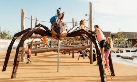 Natural playgrounds offer restorative play: young kids climbing on spider-like playground installation on sand next to a pond installation