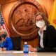 New Mexico supports tribal adoption: 2 women wearing masks and business clothessit in front of a wood wall with large, round, carved wood New Mexico State Seal with United States and New Mexico state flags to each side.