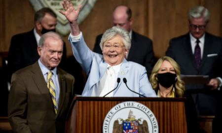 Transgender youth Alabama: Older, gray-haired woman wearing light blue suit raises right arm behind brown podium with Alabama state seal surrounded by men in dark suits