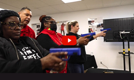 Permitless Carry Georgia: Inside a white-walled classroom, t hree women hold blue training guns at shoulder height aiming in front of themselves while male instructor stands behind them.