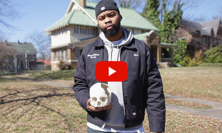 Gun violence survivor campaign: Black man in jeans, a gray winter jacket and navy baseball cap on backwards stands on sidewalk in front of s 2-story oldr home holding a white human skull with large section of bone missing on right side.