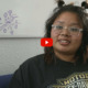 New Mexico Homeless youth: Young woman with dark hair and clear-framed glasses wearing black sweatshirt with Harlet-Davidson graphic in gold sits in couch speaking into camera