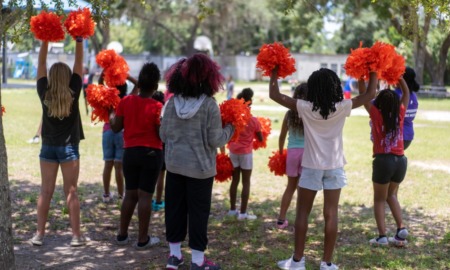 YMCA summer program: Several children in red t-sirts with red pompoms in the air stand facing a grassy field with backs to camera