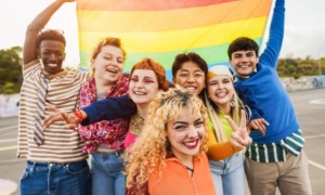 LGBTQ+ community support: Young diverse people having fun holding LGBT rainbow flag outdoor - Focus on center blond girl