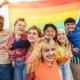 LGBTQ+ community support: Young diverse people having fun holding LGBT rainbow flag outdoor - Focus on center blond girl