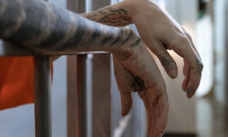 Girl vs. boy juvenile incarceration: A person's tattooed forearms and hands dangle between cell bars.