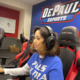 Esports: Young woman wearing headset sits at large computer screen in gaming room with multiple computers.