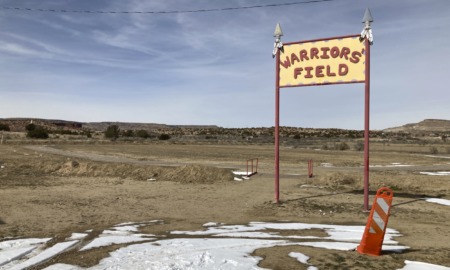 Native American Education: Tellow wood sign on two red poles with red letters reading "Warriors' Field" stands alone in a desert landscape.