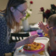 AI Flags Disabled Parents: Blonde woman with dark-framed glasses in print blouse holds spoon feeding infant in high chair from bowl of food.