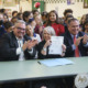 New Mexico: Large group of children stand behind two men and a woman, all in dark business suits sitting at mint green table, the men clapping and the woman holding up a paper in her hand