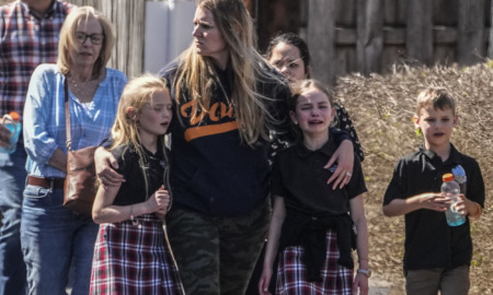 Gun violence: 3 adults , one woman hugging two of the three children, walk with crying children outside during the day.