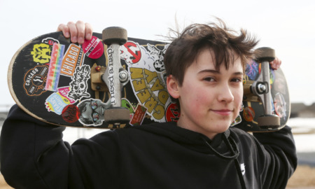 Transgender treatment for kids: Young peron with shprt dark hair on black sweatshirt stands holding colorful skateboard behine their head with both hands.