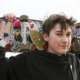 Transgender treatment for kids: Young peron with shprt dark hair on black sweatshirt stands holding colorful skateboard behine their head with both hands.