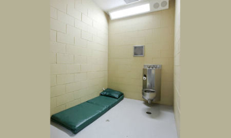 Juvenile solitary: Youth in dark, loose pants & shirt stands with back to camera in small white cell with built-in cement bed.