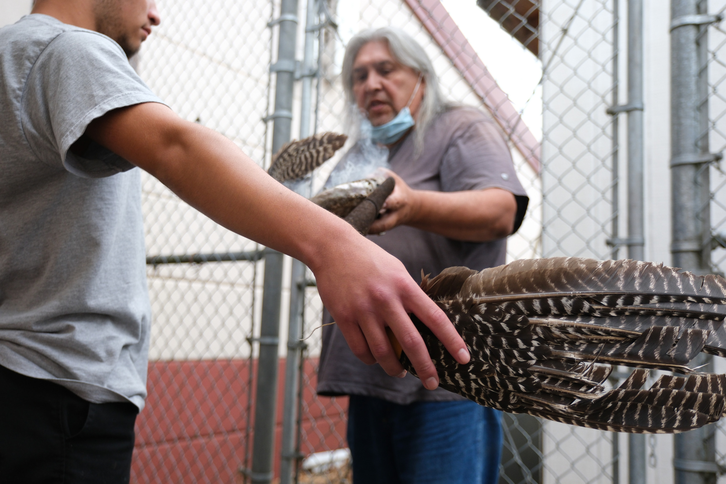 Native American rituals: Native American elder man with long gray hair in background holding smoking sage with arm of youth holding two eagle feathers in foreground