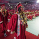 Teen Native American girl in red graduation cap and gown walks toward front of auditorium with seated fellow graduates in the background