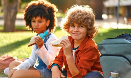 Free summer lunch: Two young children sit cross-legged on the grass eating sandwiches.