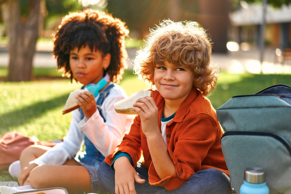 Free summer lunch: Two young children sit cross-legged on the grass eating sandwiches.