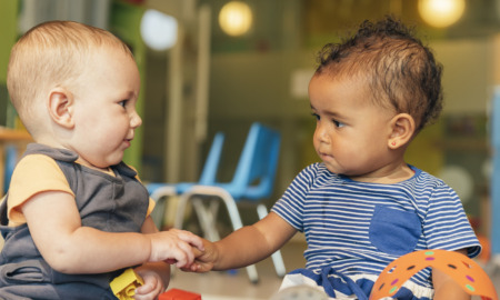 Home-based childcare business: Two young infants sit on floor side-by-side holding hands looking into each others eyes.