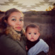 Wiyot Foster Youth Housing: Woman with long brown hair in brown winter coat with large fur collar stands outside in a filed of brown grass looking into camera holding infant draped in light blue blanket in her arms