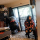 Washington homelessness and foster care: Young woman with long, dark, curly hair in red and black plaid jacket, black top and ripped jeans, sits in office chair in room with large sliding glass door.