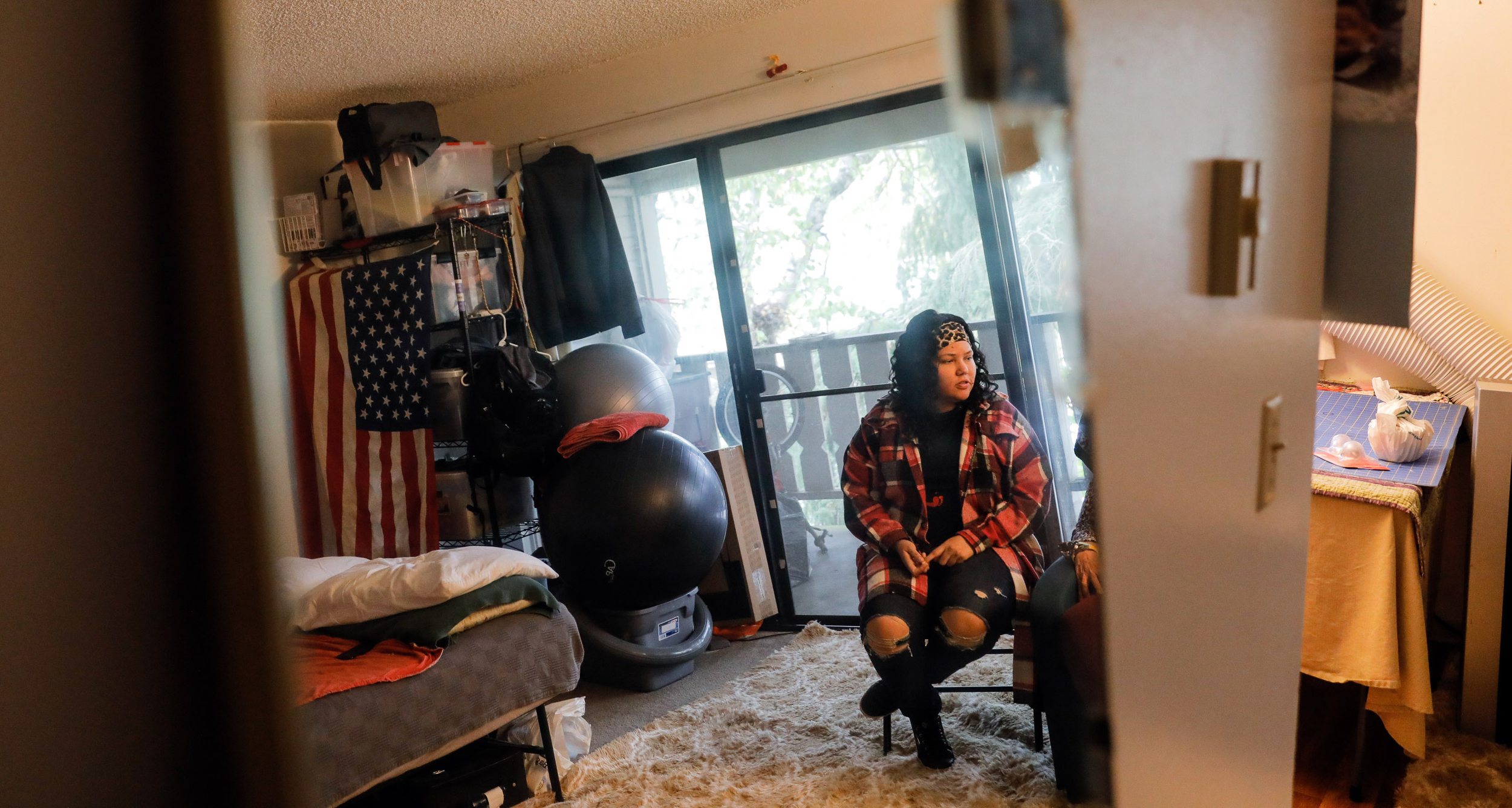 Washington homelessness and foster care: Young woman with long, dark, curly hair in red and black plaid jacket, black top and ripped jeans, sits in office chair in room with large sliding glass door.