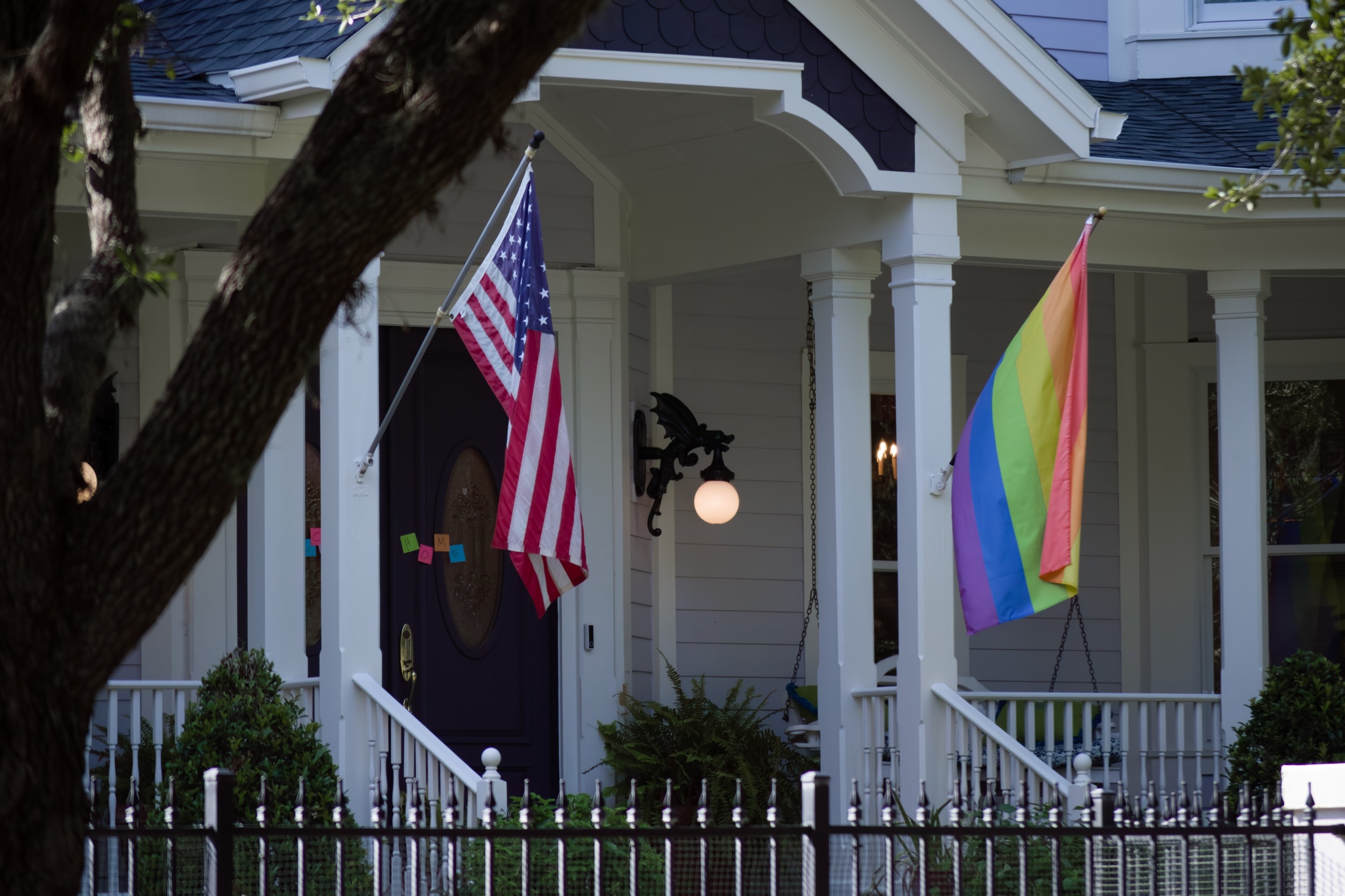 LGBTQ+ safe homes: Traditional style architecture home closeup of white front porch with black door, stairs and two pillars — one with American flag the other with a rainbow flag.