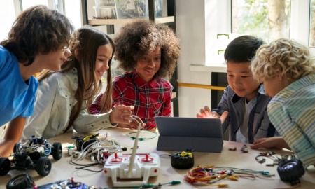 Vermont afterschool program: Group of diverse elementary students gather around a table full of robotic parts leaning in to look at a laptop screen