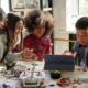 Vermont afterschool program: Group of diverse elementary students gather around a table full of robotic parts leaning in to look at a laptop screen