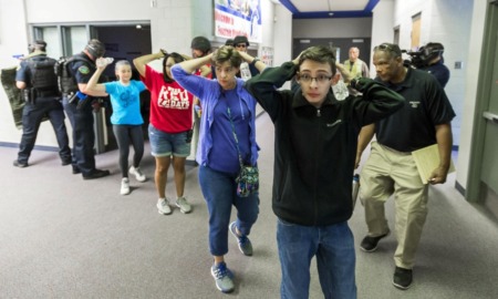 School shootings drill: Line of several teens with hands up behind heads stand in side building hallway protected by armed police