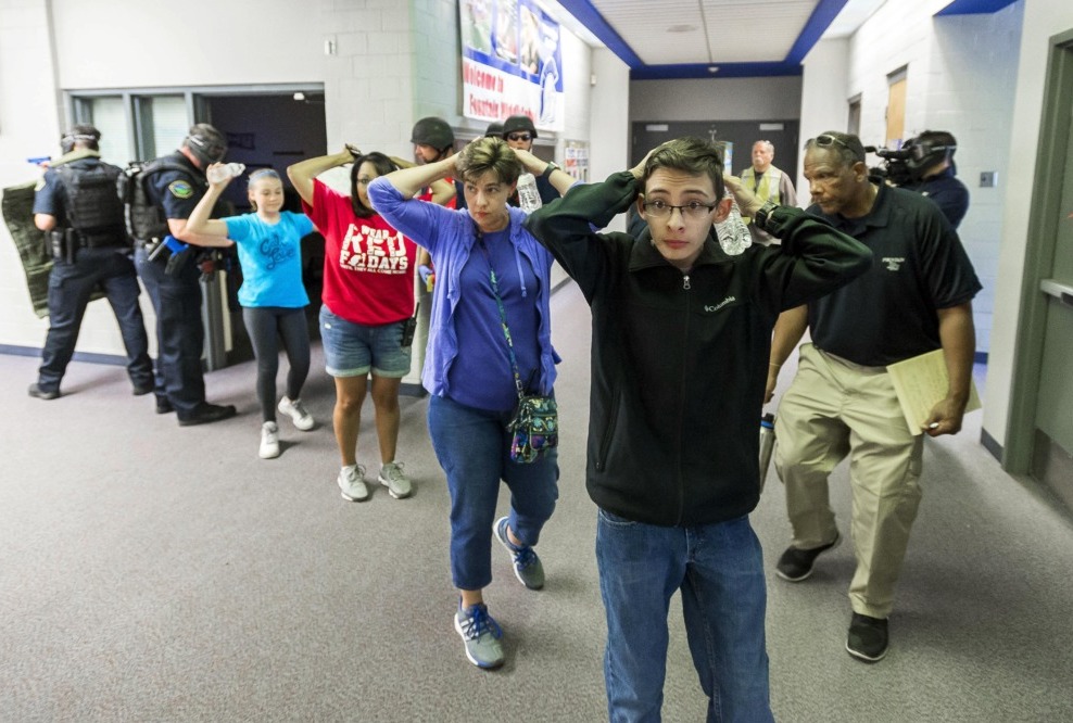 School shootings drill: Line of several teens with hands up behind heads stand in side building hallway protected by armed police