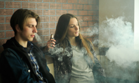 Vaping Teens & Law: Teen girl and boy sit leaning against a red brick wall vaping , heads surrounded by smoke.