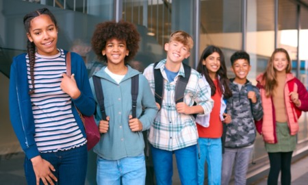 Happiness psychology: Line of happy teens stand with backpacks and book in front of a wall with floor to ceiling windows